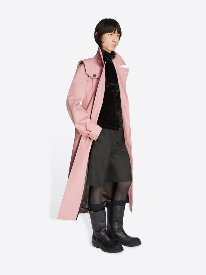 Model wearing a black turtleneck, black shorts, black leather boots and a rose colored cotton trench coat on a white background