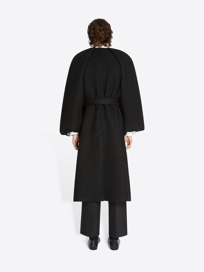 Backside of a person wearing black pants, black leather boots and a midnight colored, constructed wool coat on a white background