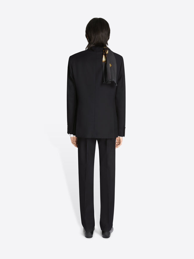 Backside of a person wearing a black, woolen, slim fit tuxedo and dark shoes on a white background