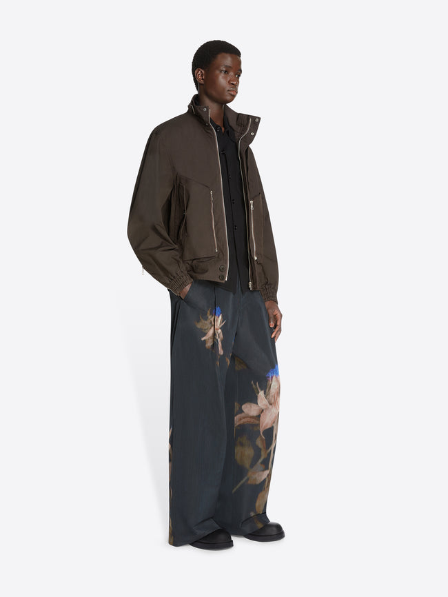 Model wearing a black shirt, brown overdyed shell jacket and black floral pants on a white background