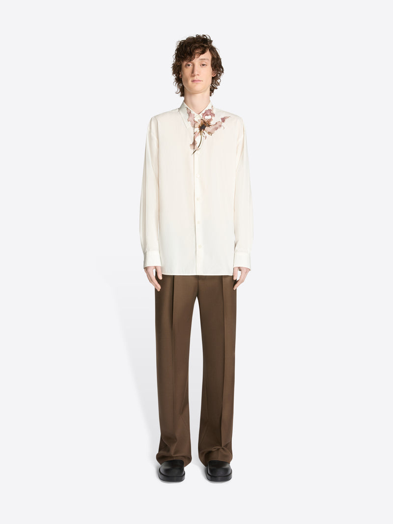 Model wearing a white, loose cotton shirt with a pink floral print and brown pants on a white background