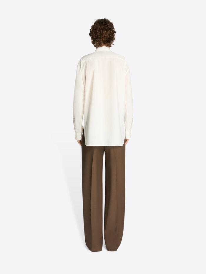 Backside of a model wearing a white, loose cotton shirt and brown pants on a white background