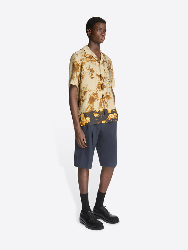 Model wearing a beige, floral print, boxy shirt and navy shorts on a white background