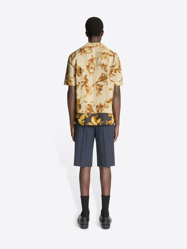 Backside of a model wearing a beige, floral print, boxy shirt and navy shorts on a white background