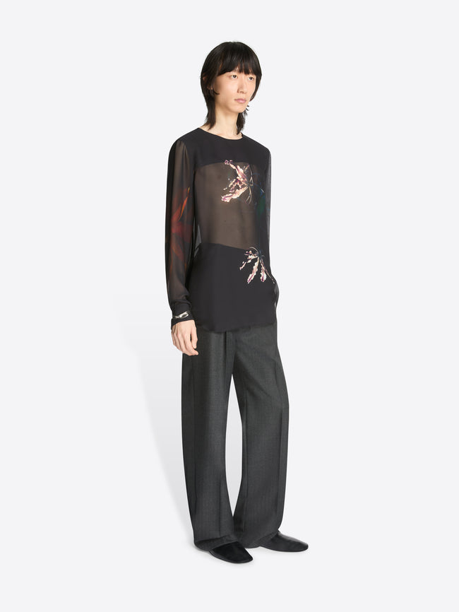 Model wearing a black, sheer collarless shirt with floral pattern and grey pants on a white background