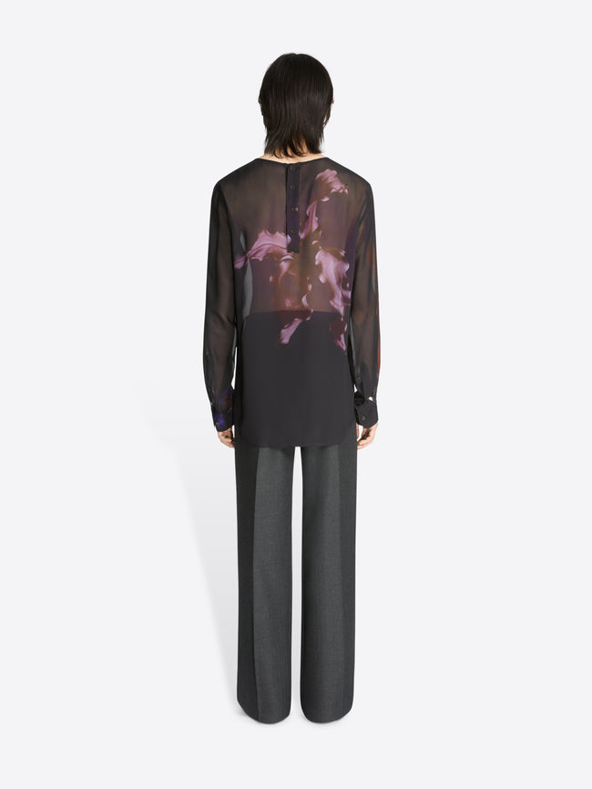Backside of a model wearing a black, sheer collarless shirt with floral pattern and grey pants on a white background