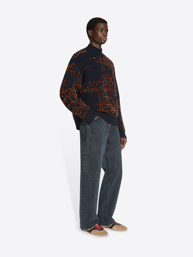 Model wearing a navy and orange patterned sweater and petrol colored loose fit jeans on a white background