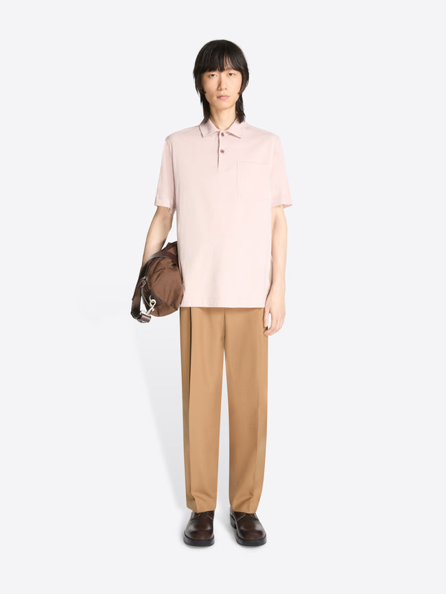 Model wearing a pale pink cotton polo shirt and beige pants while holding a brown bag on a white background
