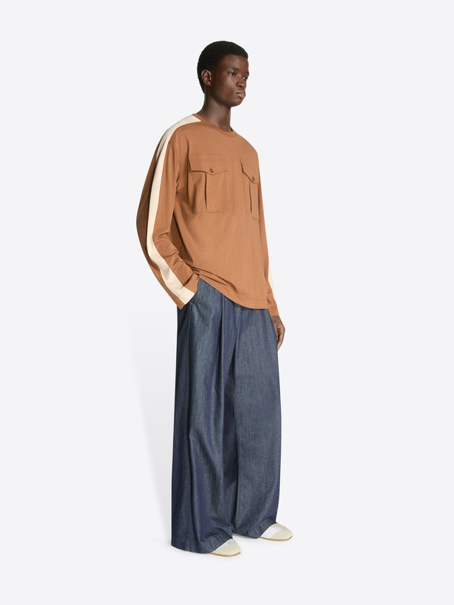 Model wearing a pair of wide, dark denim pants and a brown colored long sleeve tee with front pockets and light beige sleeve accent on a white background