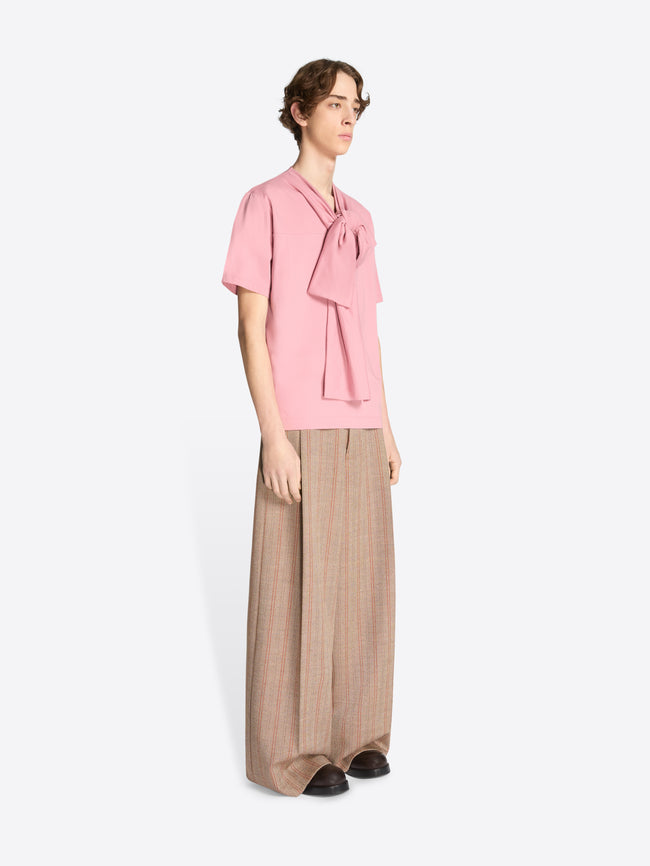 Model wearing light rose colored bow neck cotton tee and brown pants with thin red stripes on a white background
