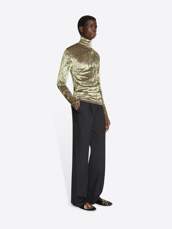 Model wearing an olive colored velvet turtleneck tee and black pants on a white background