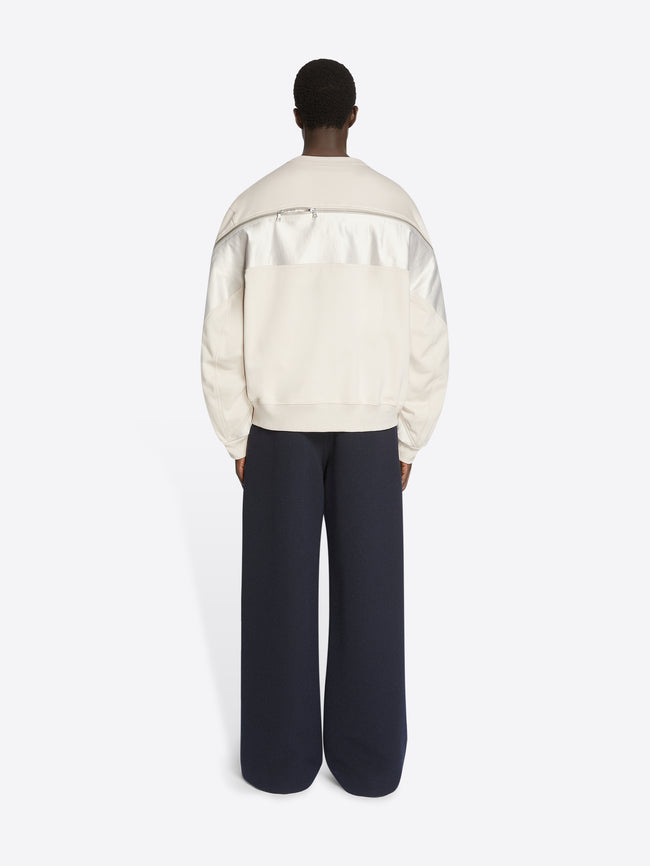 Backside of a model wearing a pearl colored sweatshirt with contrast panels and zip detailing and a pair of navy pants on a white background