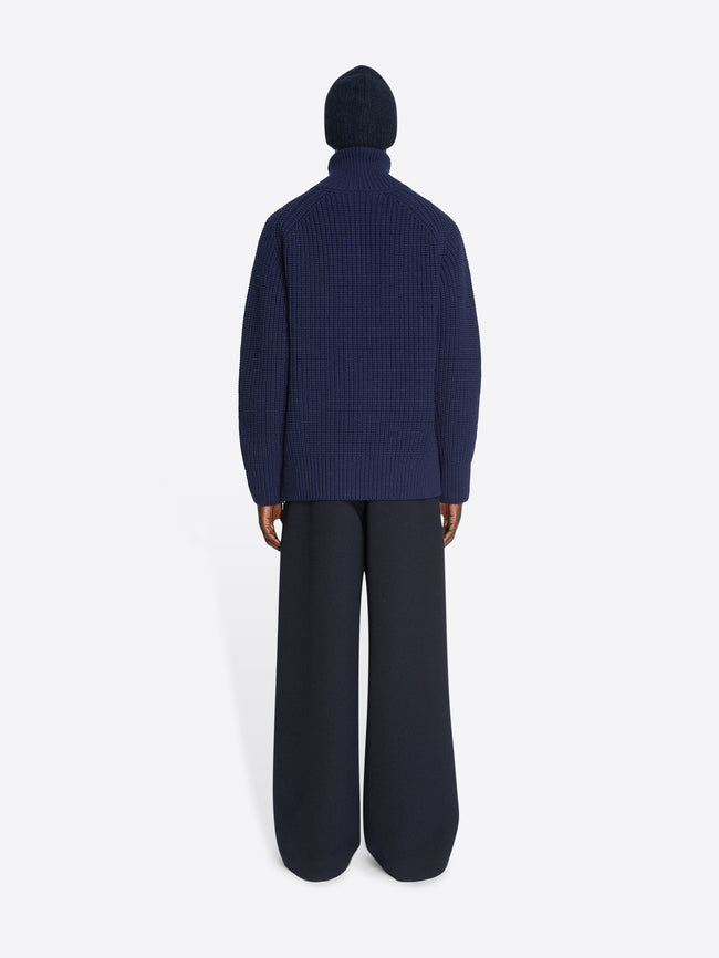 Backside of a model wearing a pair of black pants and a navy zipped wool sweater on a white background