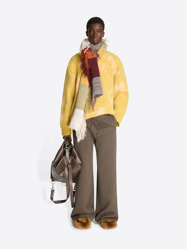 Model wearing a light yellow sweater, light brown pants and a multicolor, check pattern wool scarf with fringes on a white background