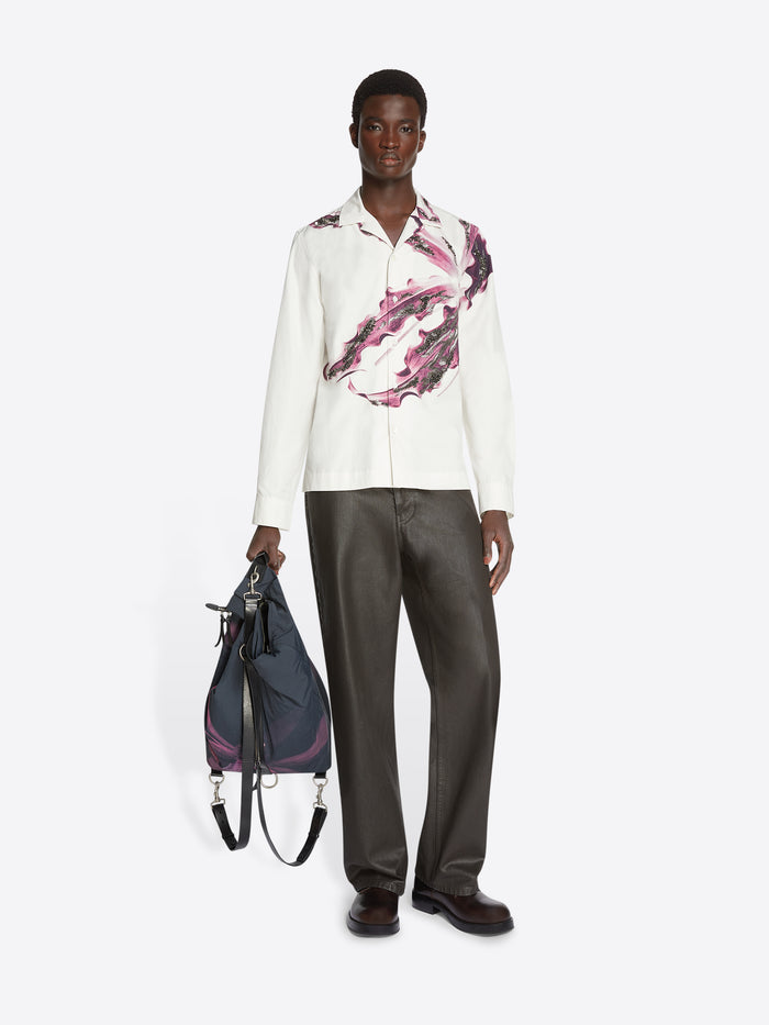 Person wearing a white shirt with a pink floral design and gray pants, holding a bordeaux black duffel bag with a floral design on a white background.