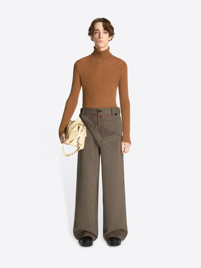 Person wearing a brown turtleneck and gray pants holding a beige bag on a white background