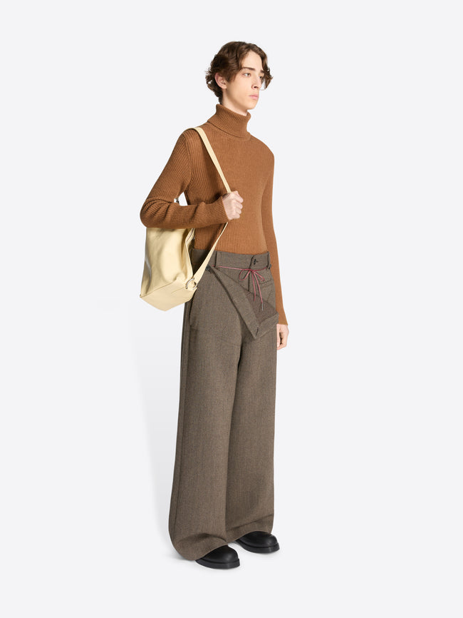Person wearing a brown turtleneck and wide-leg pants holding a sand coloured bag on a white background