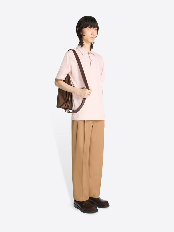 Person wearing a light pink shirt and beige pants holding a dark brown bag on a white background