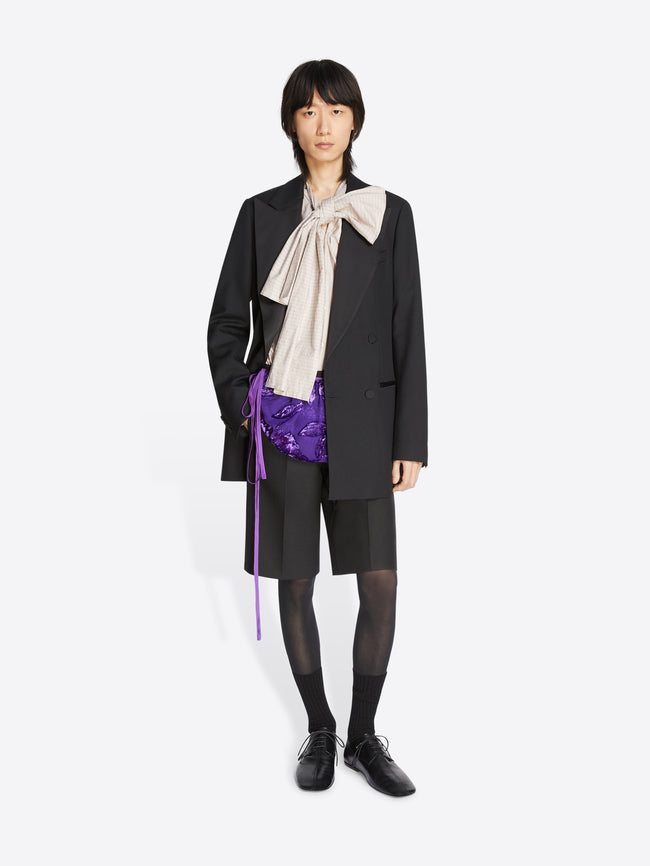 Person wearing a cream top, black blazer, jacket and leather oxford shoes on a white background