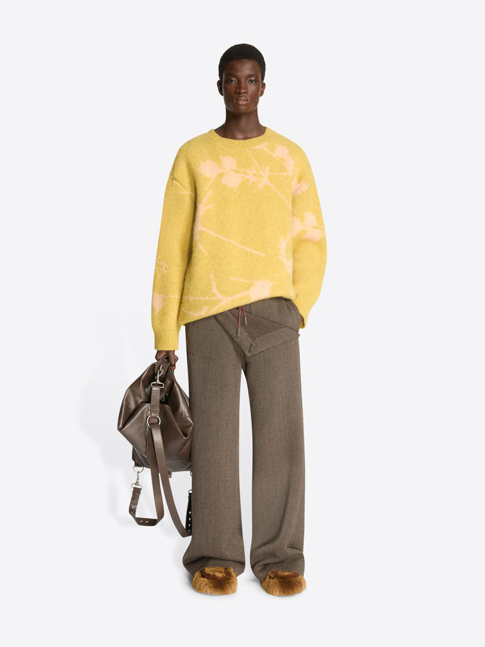 Person wearing a yellow sweater, brown pants, tan fluffy slippers and holding a brown bag on a white background