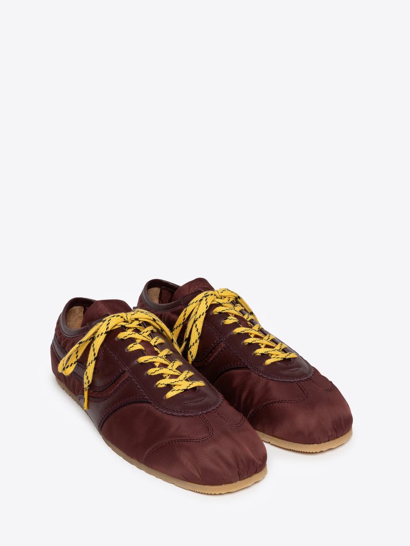 A pair of low-top sneakers in a deep burgundy fabric, featuring bright yellow braided laces and a light tan rubber sole, shown against a plain light gray background.