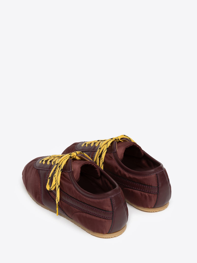 A pair of low-top sneakers in a deep burgundy satin-like material with bright yellow laces and a light tan outsole, shown from a three-quarters angle on a light gray background.