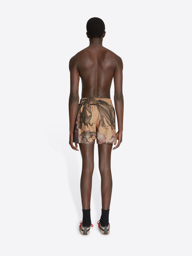 Backside of a model wearing ocra colored, floral printed swim shorts on a white background