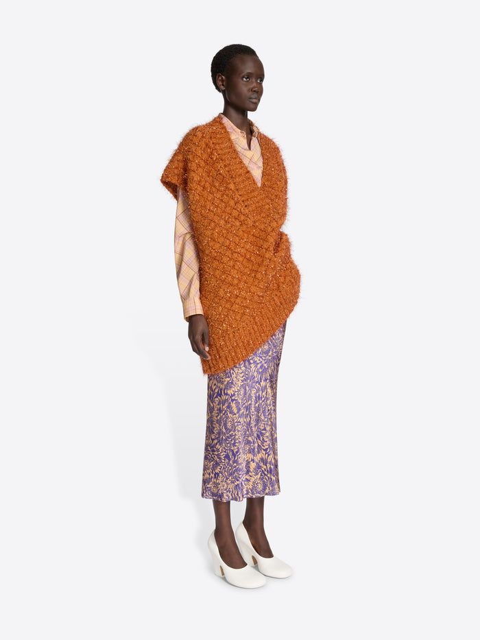 Model wearing an orange knitted poncho over a beige top and purple patterned skirt on a white background