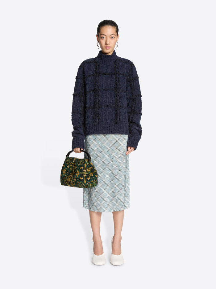 Woman wearing a dark blue textured sweater and plaid skirt, holding a patterned handbag on a white background