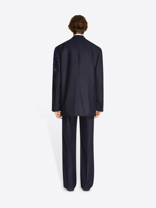 Backside of a model wearing a navy colored, floral embroidered wool blazer and matching navy embroidered pants on a white background