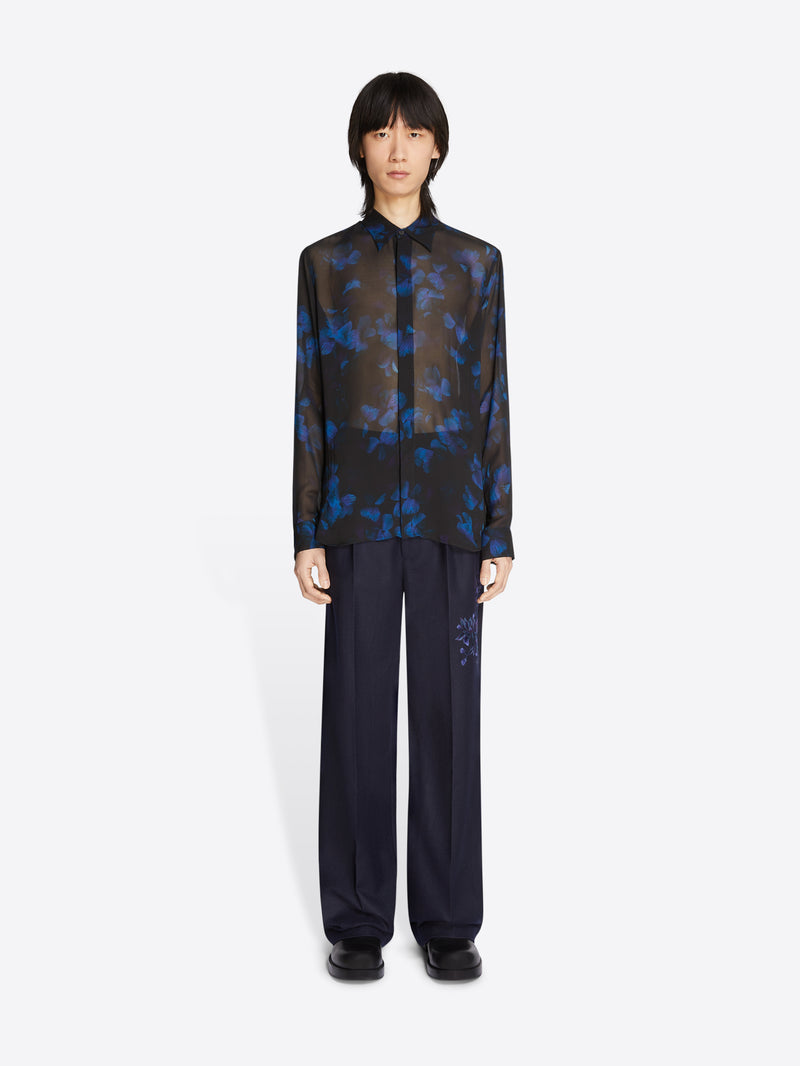 Person wearing a sheer black and blue patterned blouse with dark pants on a white background