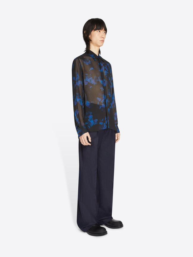 Person wearing a sheer black and blue floral shirt with wide-leg pants on a white background