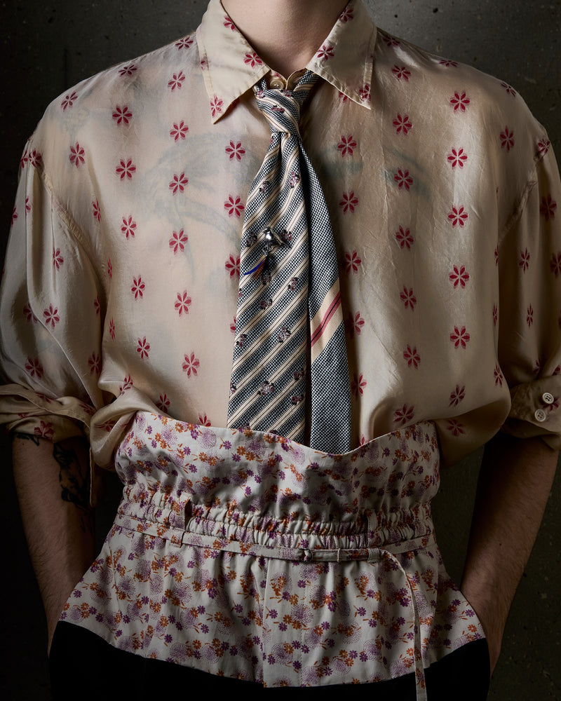 A close-up of a person wearing a beige silk-like button-up shirt with a red floral pattern, a striped blue tie, and a matching floral waist pants against a dark textured background.