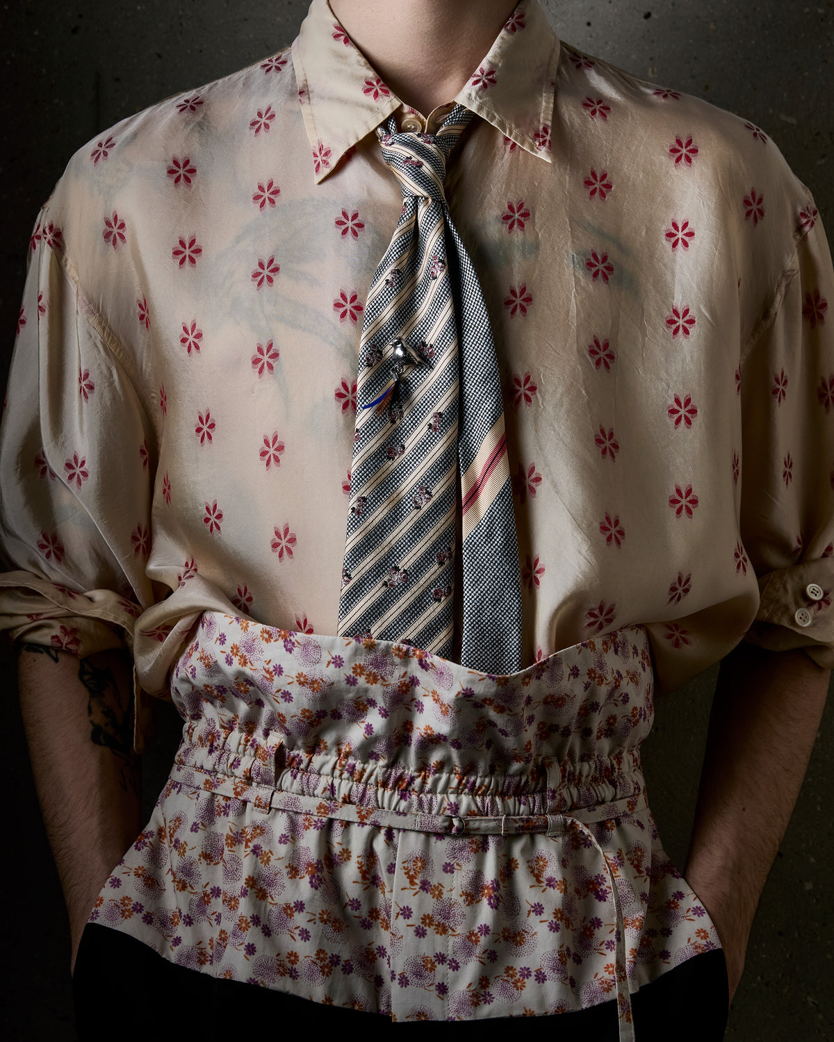 A close-up of a person wearing a beige silk-like button-up shirt with a red floral pattern, a striped blue tie, and a matching floral waist pants against a dark textured background.
