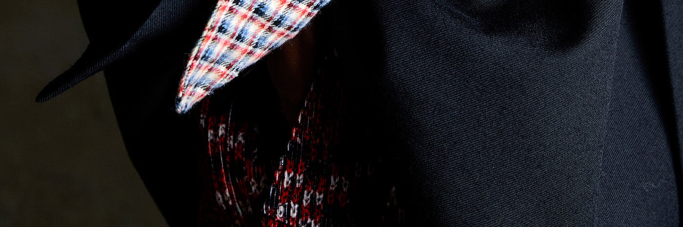 A close-up of a person wearing a black blazer over a red, white, and blue plaid patterned shirt.