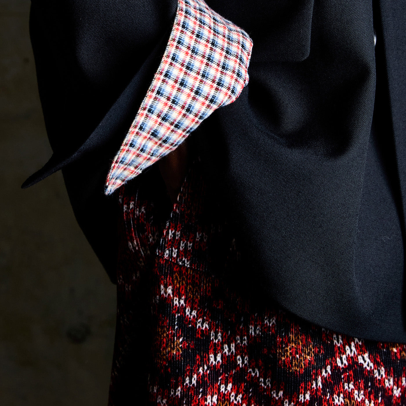 Close-up of a person wearing a black blazer over a red, black, and white patterned skirt, with a red, white, and blue plaid patterned shirt.