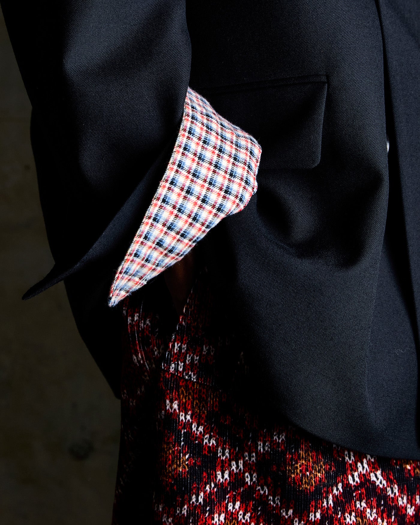 Close-up of a person wearing a black blazer over a red, black, and white patterned skirt, with a red, white, and blue plaid patterned shirt.