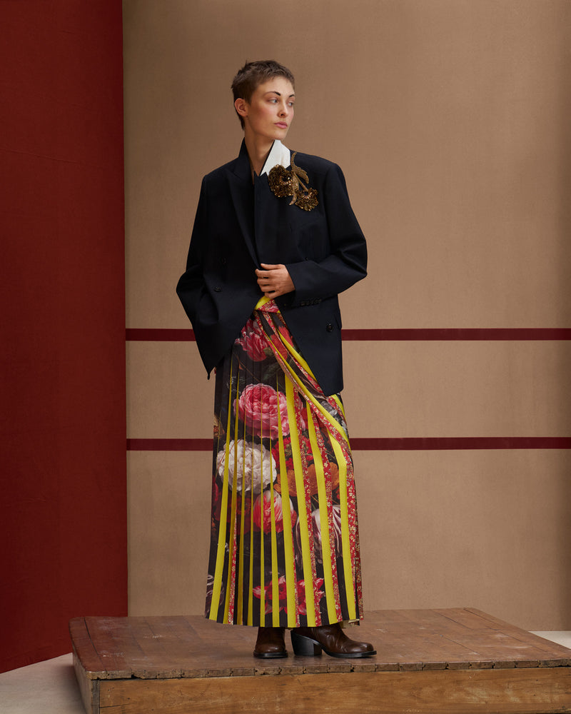 Model wearing a navy blazer with floral embellishment, a long floral and striped skirt and brown leather shoes.