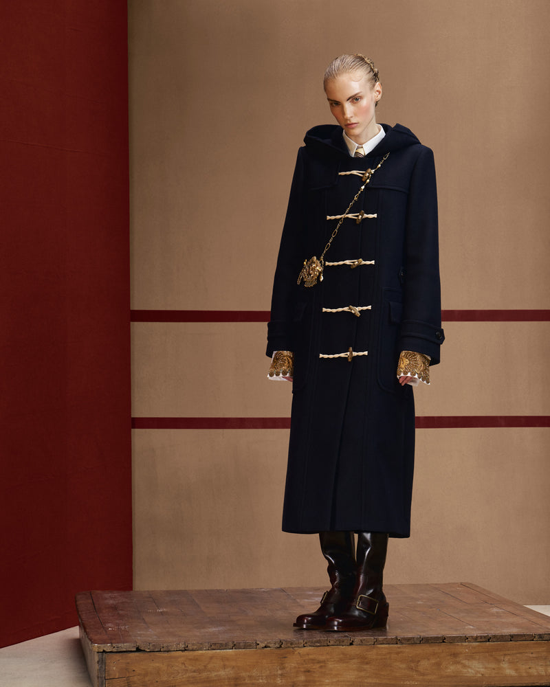 Model wearing a long navy hooded duffle coat with rope fastenings, a shirt with embellished cuffs, a gold-tone crossbody necklace and brown boots.