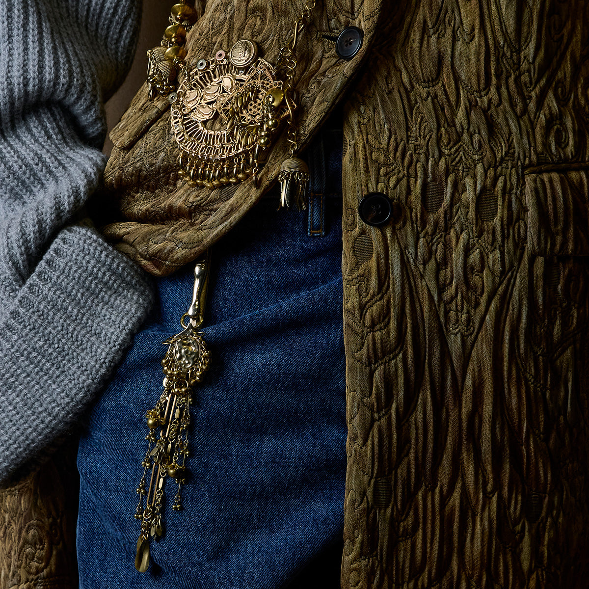 Close-up of a person wearing a chunky textured brown jacket, blue denim jeans, a gray knit sweater, and an ornate gold-toned statement charm.