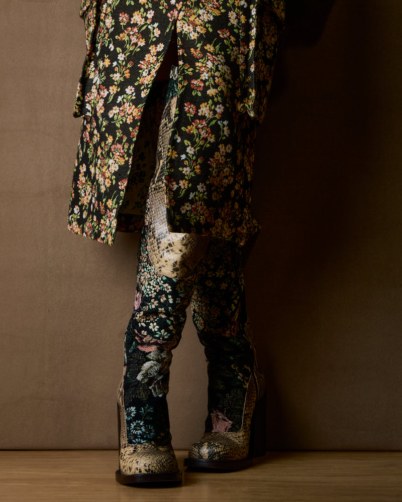 A person shown from the waist down wearing a long black floral coat, patterned boots, against a dark brown studio backdrop.