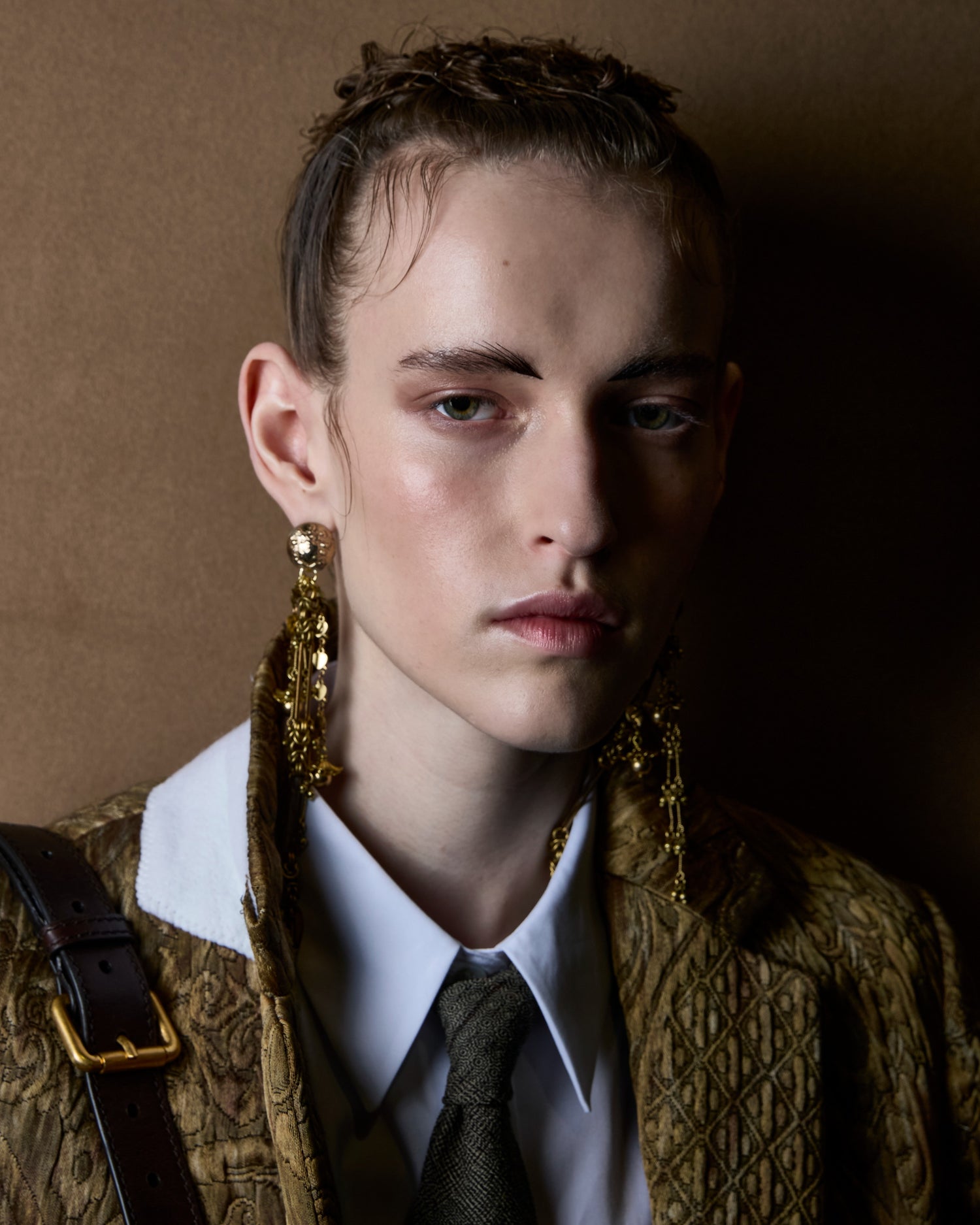 A close-up portrait of a model wearing ornate gold drop earrings, a textured patterned jacket over a white collared shirt and dark tie, against a warm brown background.