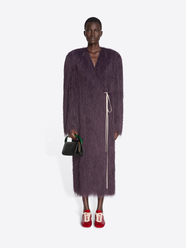 Person wearing a long dark purple coat with a green handbag on a white background - Dries Van Noten