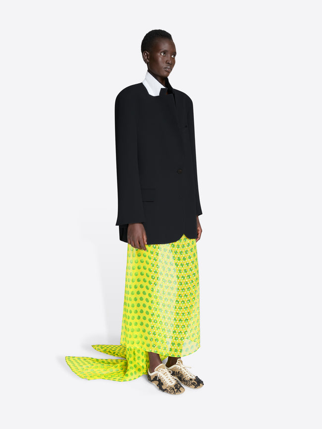 Person wearing a black suit with a bright yellow patterned skirt on a white background - Dries Van Noten