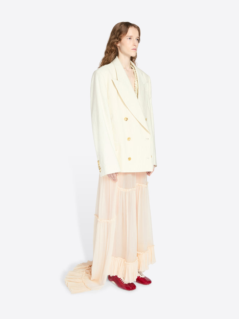 Person wearing a light-colored blazer and sheer skirt with red shoes on a white background - Dries Van Noten