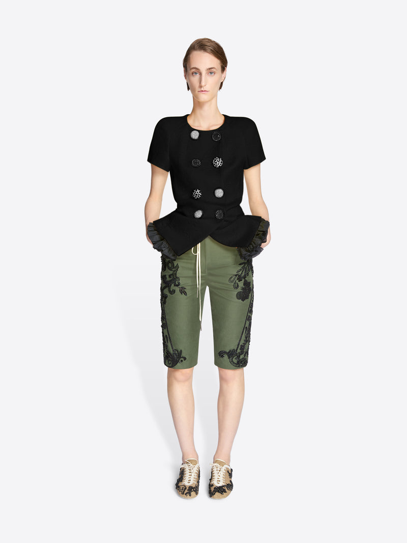 Woman wearing a black top with decorative buttons and green pants with floral patterns on a white background - Dries Van Noten