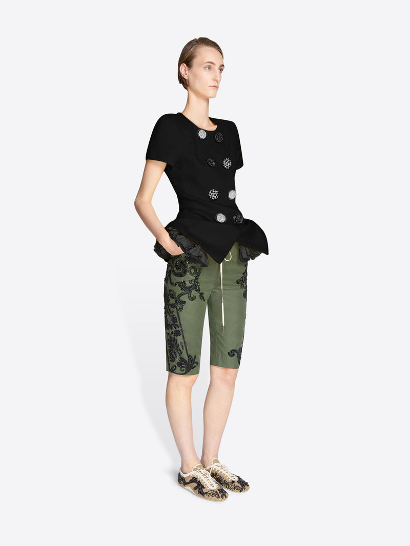 Person wearing a black top with silver buttons and green camouflage shorts on a white background - Dries Van Noten