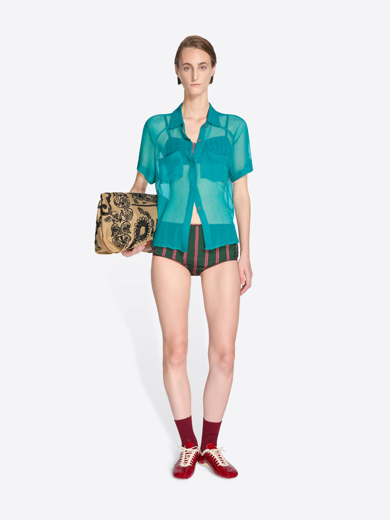Woman wearing a green sheer blouse and black shorts, holding a patterned clutch on a white background - Dries Van Noten