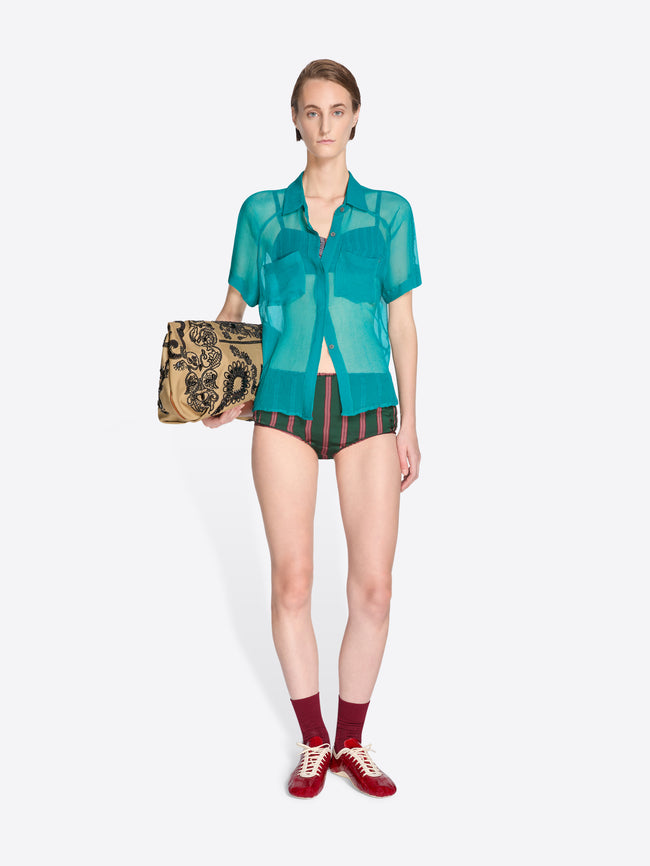 Woman wearing a green sheer blouse and black shorts, holding a patterned clutch on a white background - Dries Van Noten