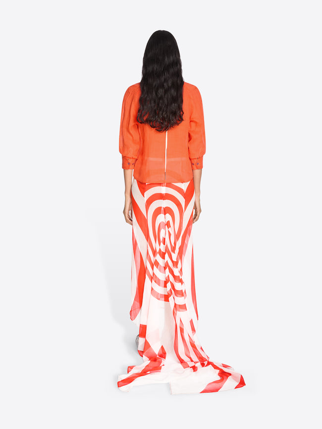 Woman wearing a bright orange top and a long, patterned skirt with white and red design on a white background - Dries Van Noten